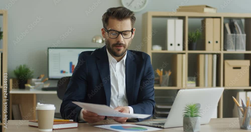 Caucasian handsome young man in glasses and official style having a business videochat on the laptop in his office and demonstratingsome charts with data tothe web cam. Caucasian handsome young man in glasses and official style having a business videochat on the laptop in his office and demonstratingsome charts with data tothe web cam.
