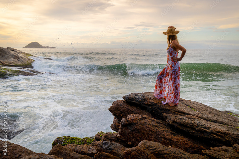 Beautiful slender woman with stylish dress standing on a beach with the waves coming in and the wider seascape at sunrise Beautiful slender woman with stylish dress standing on a beach with the waves coming in and the wider seascape at sunrise