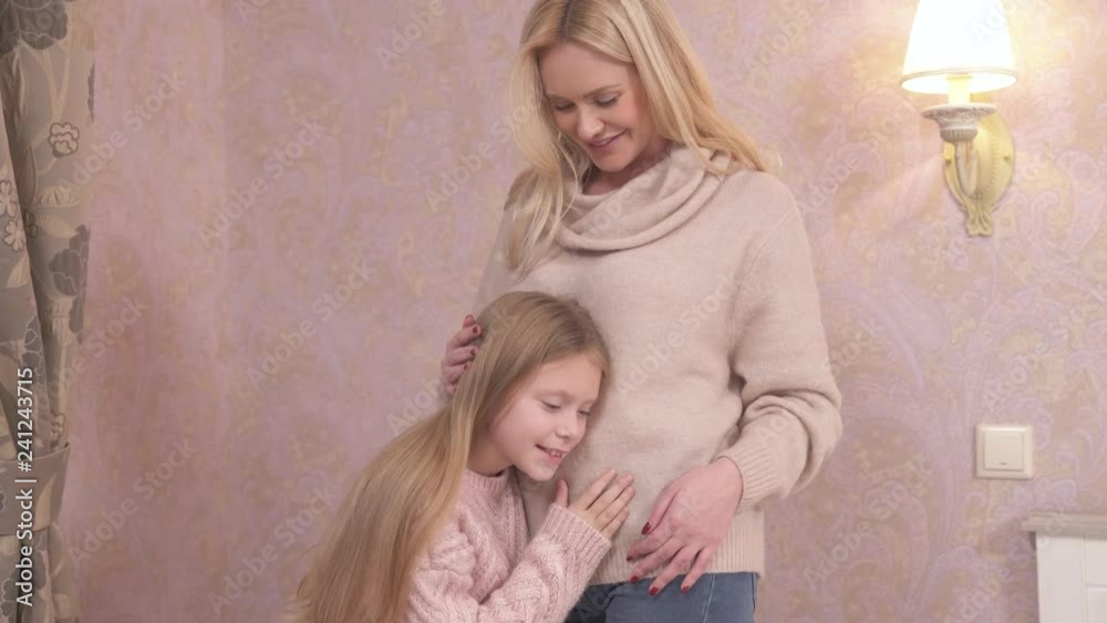 Fair-haired woman and girl are standing in the room. The girl is stroking woman's belly and putting her ear to it listening. They are happy and smiling Fair-haired woman and girl are standing in the room. The girl is stroking woman's belly and putting her ear to it listening. They are happy and smiling