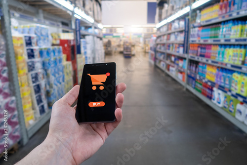 Hand holding mobile phone on Supermarket blur background. Shopping basket on a mobile phone screen. 