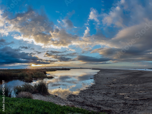 Glowing sun rays peek from horizon clouds to illuminate the smooth and reflective surface of pond at lower tide of ocean lagoon.