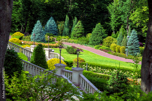 Stairs with stone railings balusters and iron lanterns on the background of the park with a luxurious landscape design walking path for walking, green lawns and a variety of bushes.