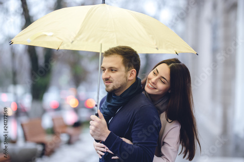 Happy romantic couple, guy and his girlfriend dressed in casual clothes are hugging under the umbrella and look at each other on the street in the rain.