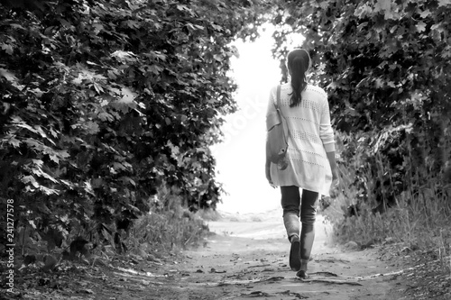 The girl walks along the path in the summer woods to the light in a white jacket and jeans.