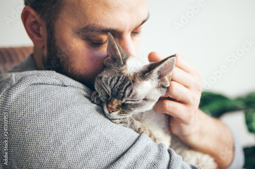 Portrait of happy cat with close eyes and young beard man snuggling. Handsome young man is hugging and cuddling his cute color point Devon Rex kitten. Domestic pets. Kitty likes attention and purrs