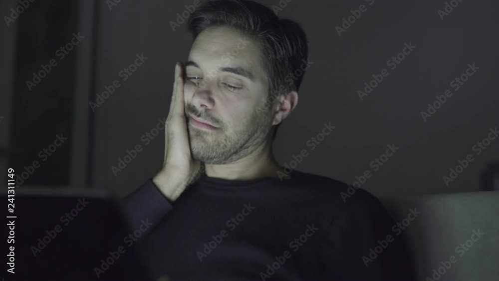 Exhausted man rubbing face and using digital tablet at night. Tired sleepy young man using tablet computer in dark room. Working late concept Exhausted man rubbing face and using digital tablet at night. Tired sleepy young man using tablet computer in dark room. Working late concept