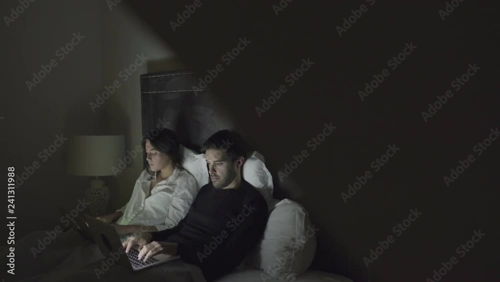 Focused couple using laptops in bedroom at night. Selective focus of concentrated young man and woman working with laptops together in bed. Technology concept Focused couple using laptops in bedroom at night. Selective focus of concentrated young man and woman working with laptops together in bed. Technology concept