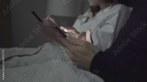 Cropped shot of couple using smartphones in bed. Close-up partial view of young man and woman lying in bed and using mobile phones at night. Technology concept