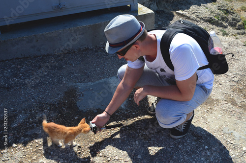 Caucasian man with a backpack crouched down and holding an action camera in his outstretched hand. Red kitten sniffs the camera