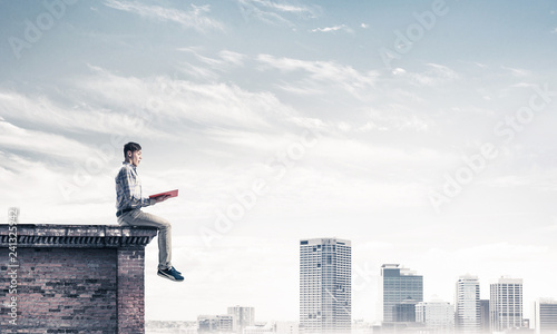Man on roof edge reading book and cityscape at background