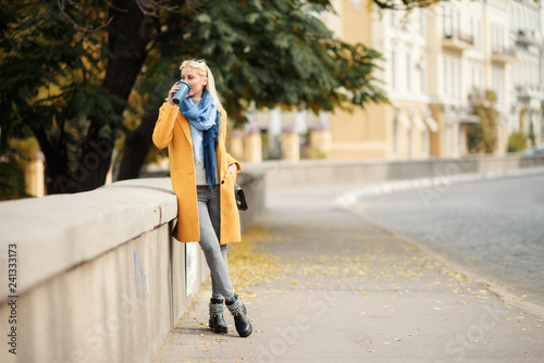 Stylish happy young blond woman in bright yellow coat holds coffee to go. Walking on the street. Portrait of a young cheerful woman at work break. Lifestyle concept 