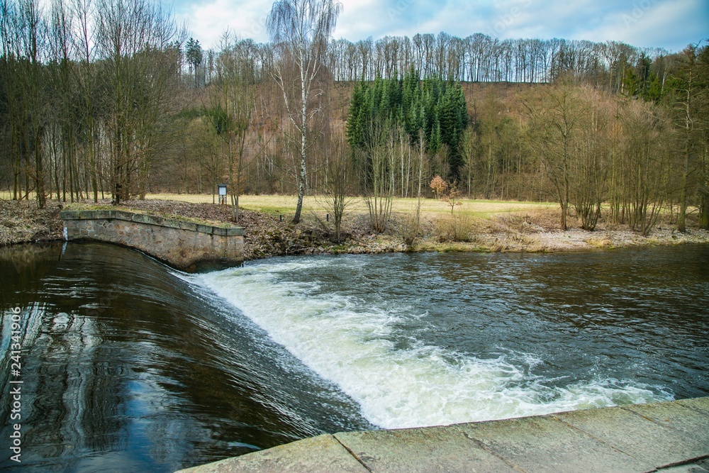 View of spring nature landscape and a dangerous weir at river Upa, dark water, white riffles, the Czech Republic, Europe, blue sky, white clouds, trees standing along river, hill in distance