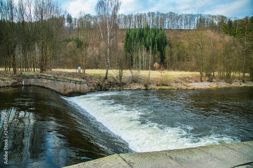Obraz na płótnie View of spring nature landscape and a dangerous weir at river Upa, dark water, white riffles, the Czech Republic, Europe, blue sky, white clouds, trees standing along river, hill in distance w salonie