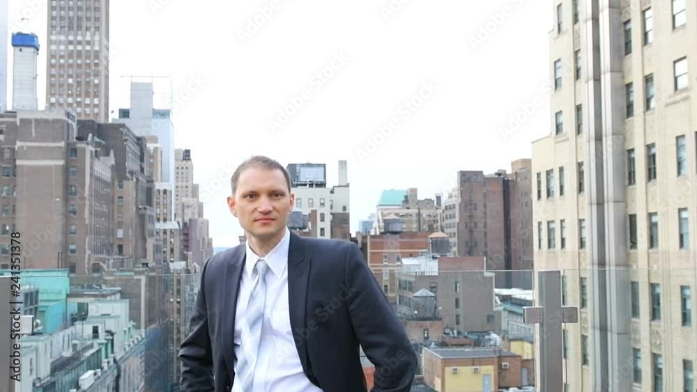 Slow motion of happy, smiling young businessman, man looking at camera with business suit on top of rooftop, roof with urban aerial view on skyscrapers in New York City Slow motion of happy, smiling young businessman, man looking at camera with business suit on top of rooftop, roof with urban aerial view on skyscrapers in New York City