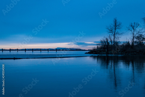 small island by the wooden piers with trees grown on it cast reflection on water surface on the river under cloudy sky at blue hour