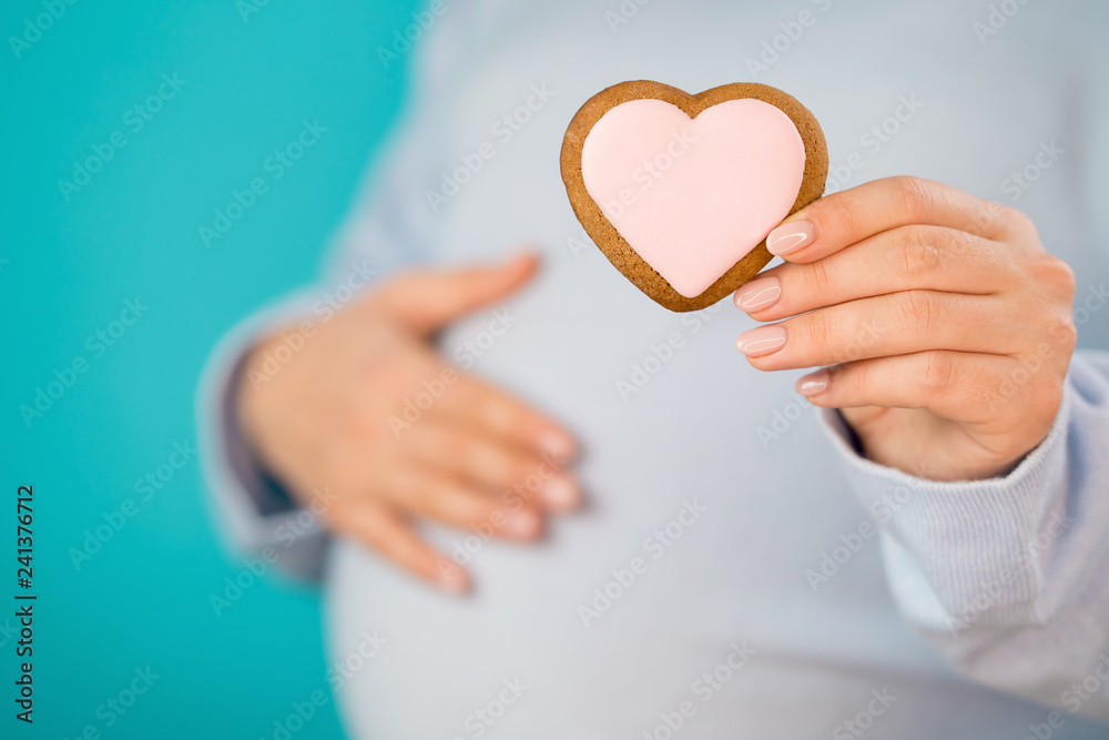 Pregnant woman holding heart-shaped cookie on tummy belly background. Young girl in blue wearing expecting baby. Maternity, motherhood, pregnancy, love concept. Pregnant woman holding heart-shaped cookie on tummy belly background. Young girl in blue wearing expecting baby. Maternity, motherhood, pregnancy, love concept.
