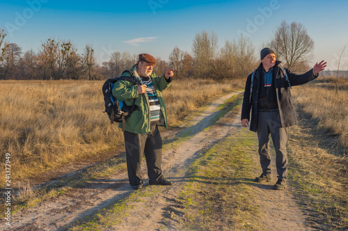Two senior hikers with backpacks discussing correct path while walking on a country road at autumnal evening