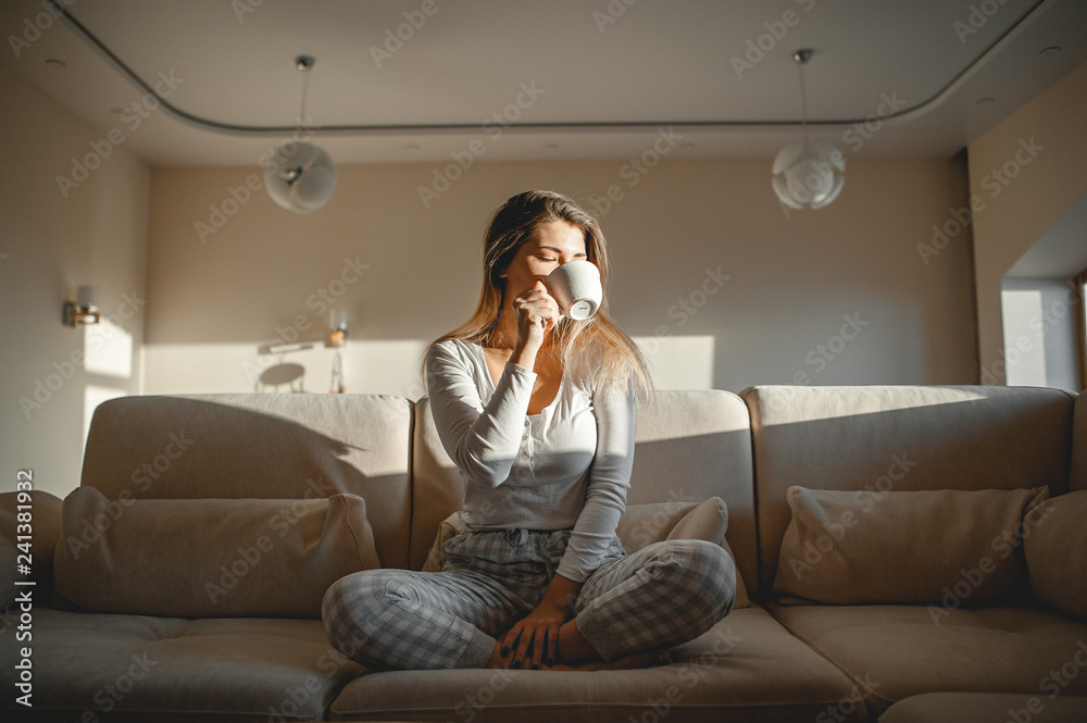 A blonde girl after breakfast sits in her pajamas in a large room on a sofa and drinks tea or coffee from a mug. A blonde girl after breakfast sits in her pajamas in a large room on a sofa and drinks tea or coffee from a mug.