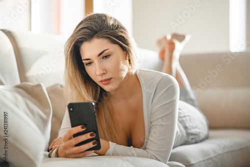 A girl in pajamas is lying on a sofa in a bright room and communicating by video-call with a friend using a wireless earpiece.