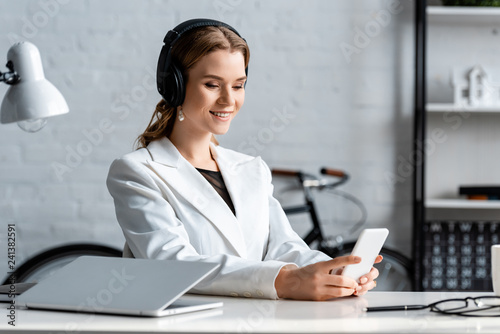 smiling businesswoman in headphones and formal wear sitting at desk and using smartphone at workplace