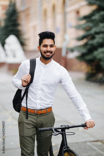 Portrait of cheerful young asian guy walking outdoors with bicycle holding backpack.