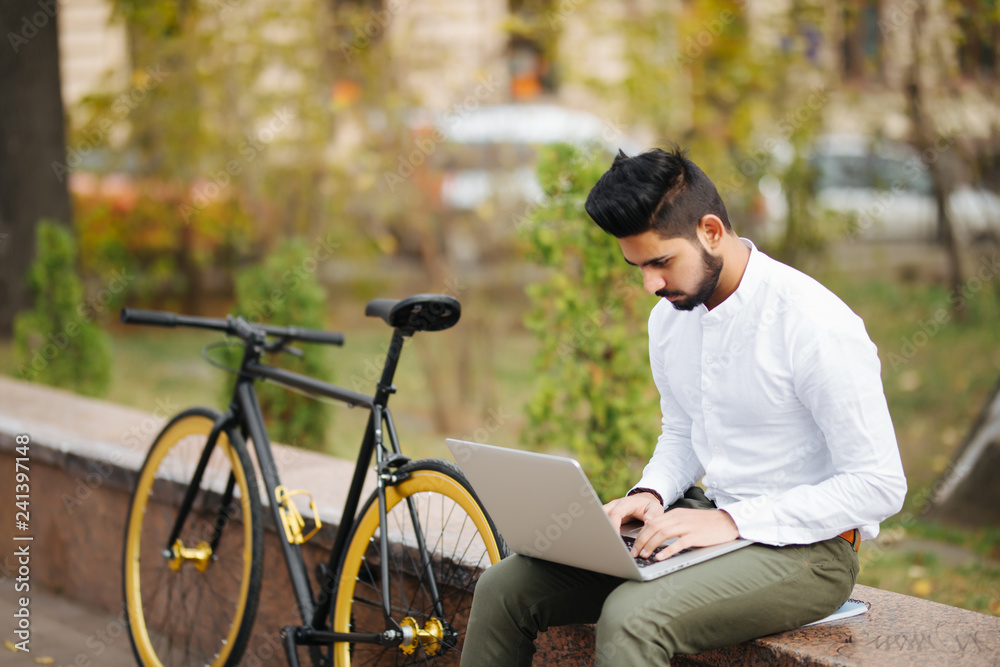 Fototapeta premium Handsome young indian businessman dressed in suit working on laptop computer while sitting outdoors near bicycle