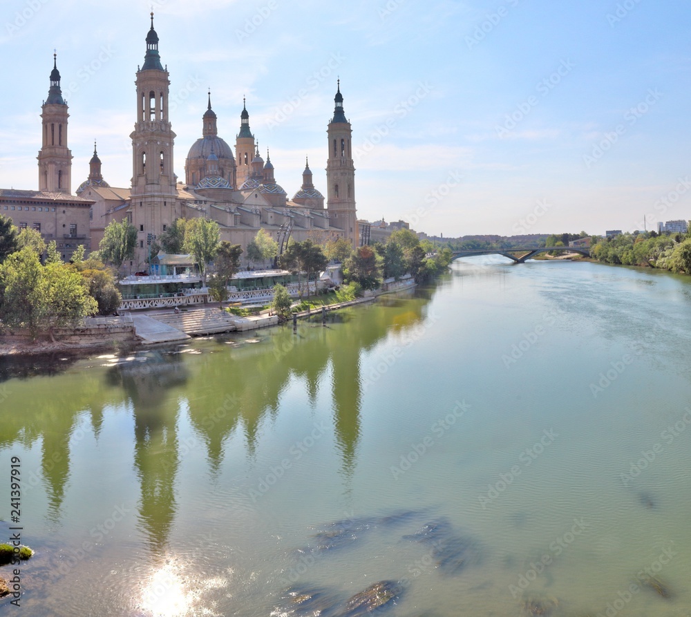 A landscape of christian Pilar Cathedral and Santiago Bridge reflecting in the Ebro river during a sunny summer day in Zaragoza, Aragon region, Spain