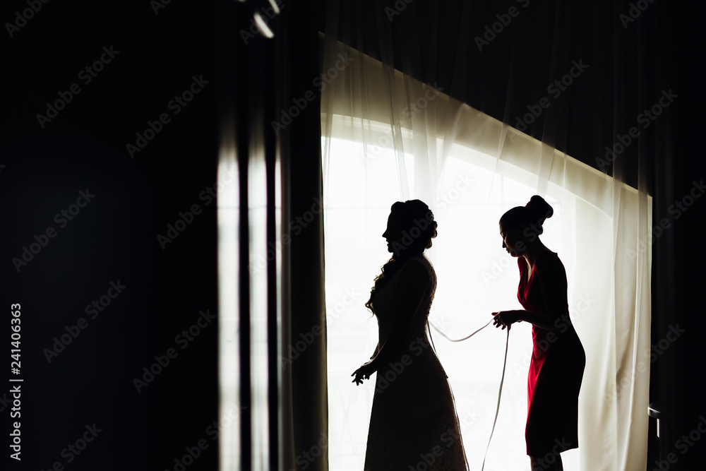 Profile of the bride and her friend who lacing corset in a wedding dress near the window
