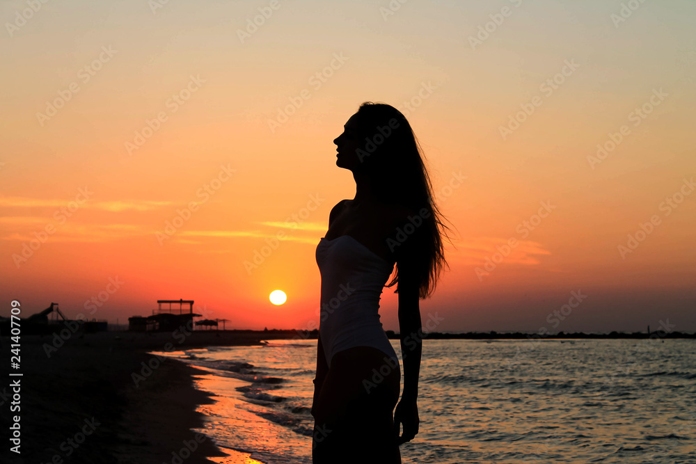 Silhouette of sexy girl in a swimsuit on a beach of the calm sea during a sunrise. Beach vacation. Magnificent sunset.