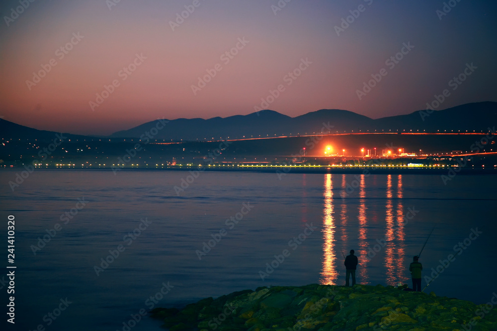 Panorama of fishermen at sunset in Gelendzhik Bay. Fishermen stand on the pier at sunset. Silhouettes, rocks, lights on the opposite side of the Bay.
