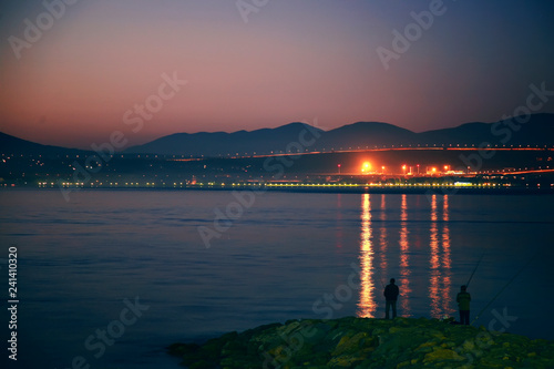 Panorama of fishermen at sunset in Gelendzhik Bay. Fishermen stand on the pier at sunset. Silhouettes, rocks, lights on the opposite side of the Bay. © Александр Трихонюк