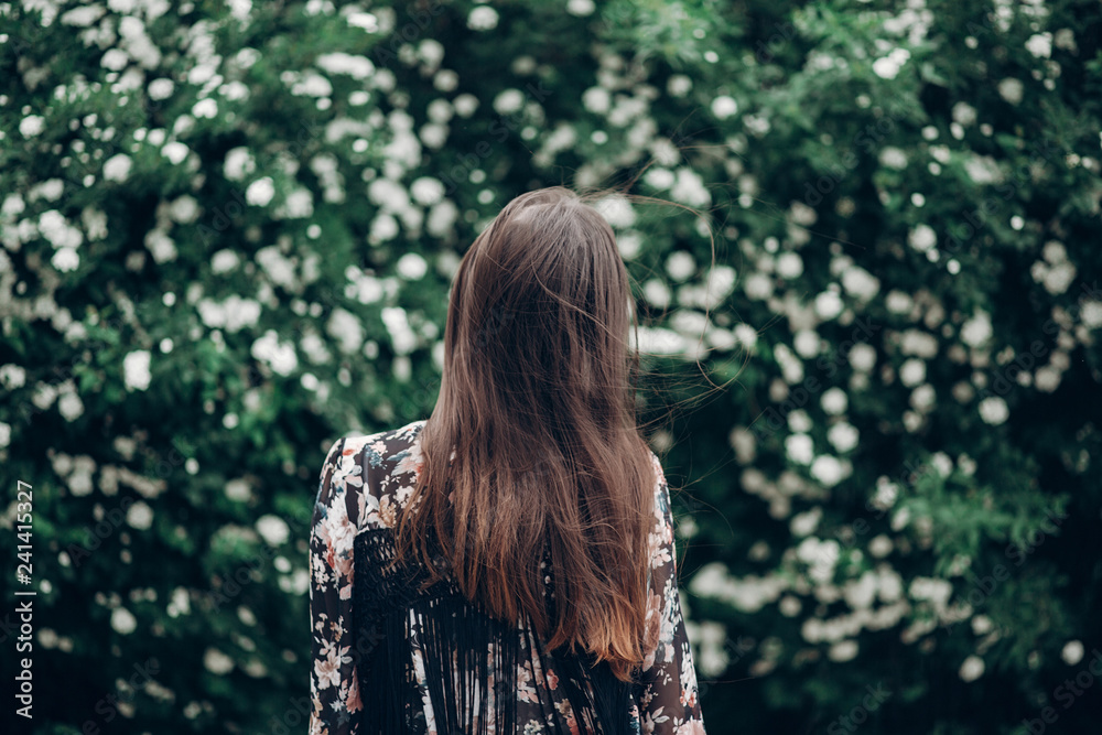 back of hipster woman posing on background of blooming bush with white flowers of spirea. boho girl waving hair in floral clothes. space for text. spring summer time