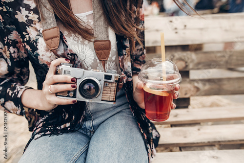 stylish hipster woman holding lemonade and old photo camera. boho girl in denim and bohemian clothes, holding cocktail sitting on wooden bench at street food festival. summertime