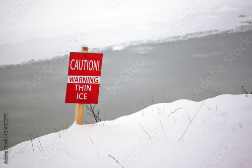 warning sign on the bank of a frozen lake with the inscription: "CAUTION! WARNING THIN ICE"
