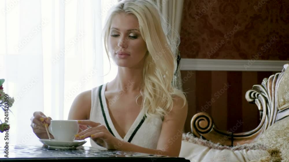 Young beautiful blonde woman in elegance white dress having breakfast and drinking a cup of tea or coffee in a luxurious living room. 4K Young beautiful blonde woman in elegance white dress having breakfast and drinking a cup of tea or coffee in a luxurious living room. 4K