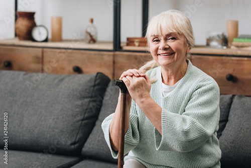 cheerful senior woman sitting on sofa and holding walking stick photo