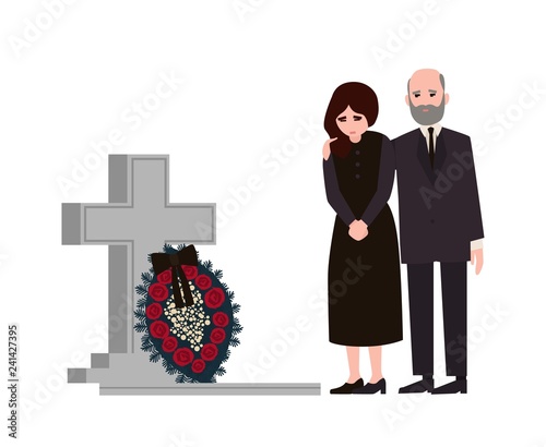 Sad man and woman dressed in mourning clothes standing near grave with tombstone and wreath