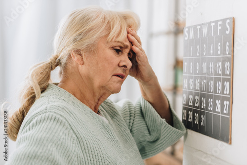 upset woman with gray hair touching head and looking at wall calendar photo