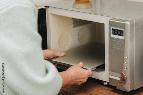 cropped view of senior woman with dementia disease putting laptop in microwave oven