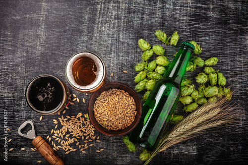 A bottle of beer with green hops, oat, wheat spikelets, opener and glasses with dark and light beer on black scratched chalk board