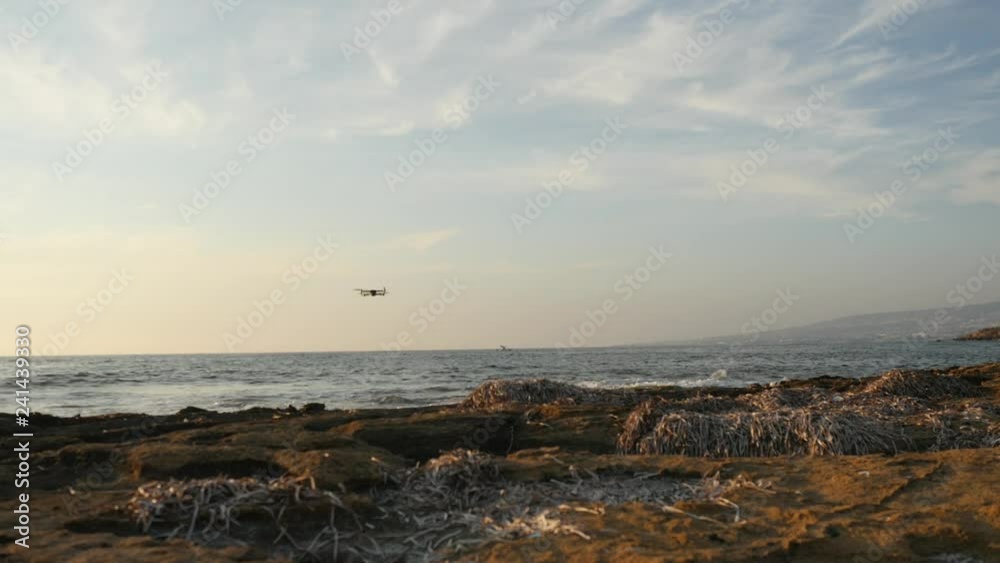 Modern quadcopter quickly flies over the water to the coast, raising the wind above the relief ground Modern quadcopter quickly flies over the water to the coast, raising the wind above the relief ground