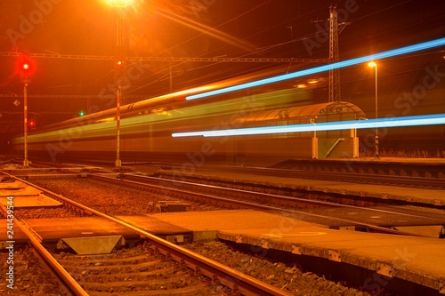 White and blue train passing a small roofless train station at night in the Czech Republic. Blurred motion train
