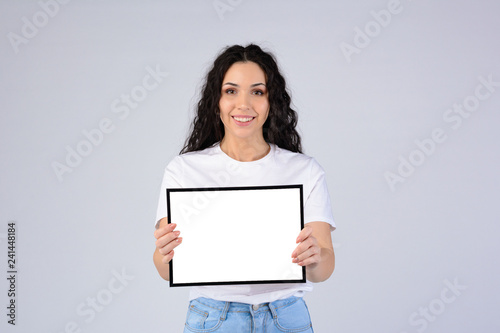Attractive curly young woman in white t-shirt holding empty blank photo frame with free copy space. Special offer. Hire me concept. Grateful letter