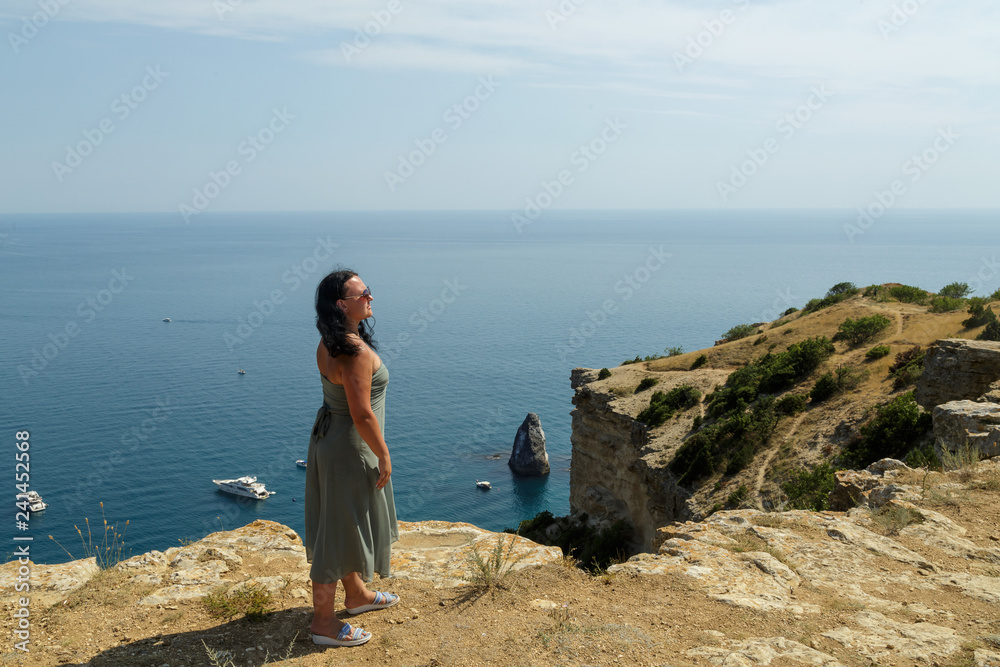 A young woman stands on the edge of a cliff and admires the view of the sea.