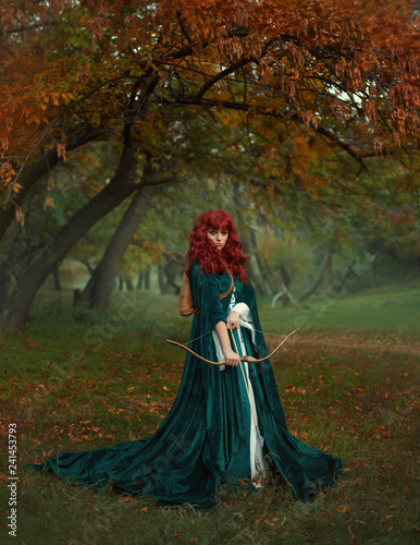 red-haired beauty in search of the victim  the legend of Robin Hood  mysterious lady in green velvet long raincoat with arrows and bow in her hands  fearless girl ready for battle in a foggy forest