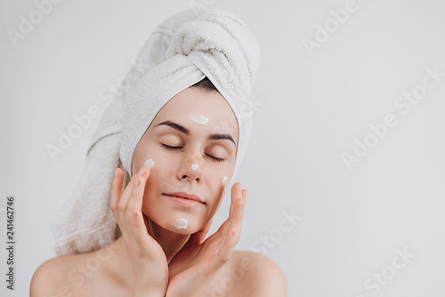 Young laughing girl with a towel on her head puts applying moisturizing cream on her face. Photo of women with flawless skin on white background. Skin care and beauty concept