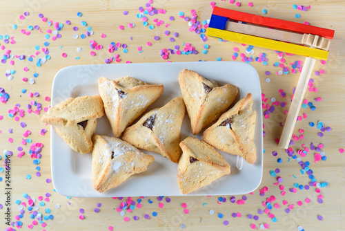 Hamantaschen cookies on white dish and wooden noisemaker or gragger on wood table. Colorful confetti background. Purim holiday celebration. Top view. photo