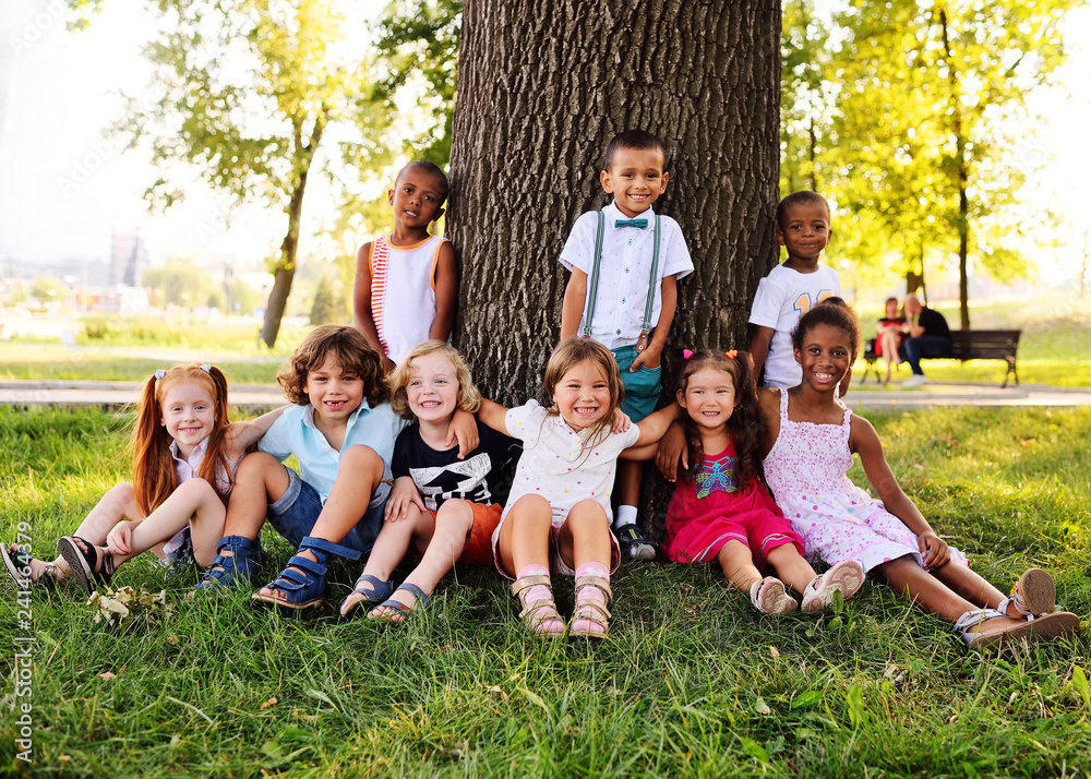 a group of children of different racial and ethnic types sit together under a big tree in the Park on the grass and smile. a group of children of different racial and ethnic types sit together under a big tree in the Park on the grass and smile.