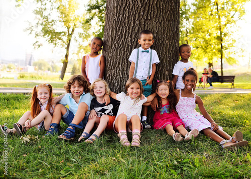 a group of children of different racial and ethnic types sit together under a big tree in the Park on the grass and smile.