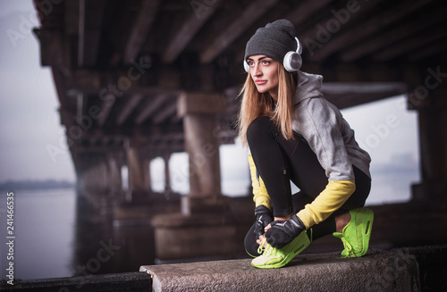 sporty woman in headphones under the bridge. Young woman resting after running in winter